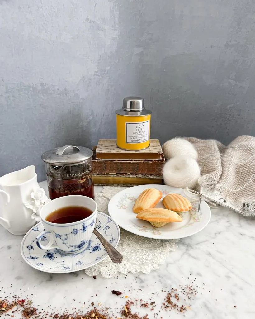 A yellow canister of tea sits on top of a tall stack of books surrounded by knitting, madeleines, a clear pot of tea, a creamer, and a blue and white teacup filled with tea.