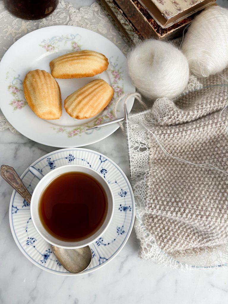 A blue and white teacup filled with tea dominates the bottom left of this photo. Around it to the top and right are a plate of madeleines, some yarn, and a handknit shawl in progress.
