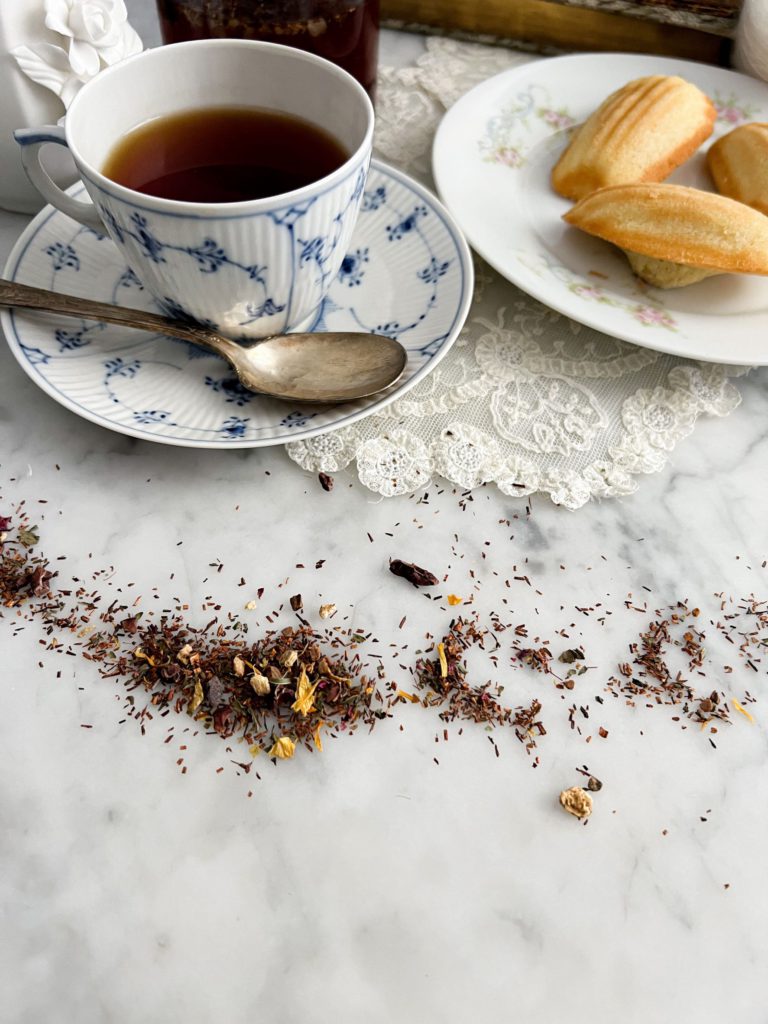 In the foreground is a spread-out pile of dried tea leaves on a white marble countertop. Behind it are a blue and white teacup full of tea and a plate of madeleines.