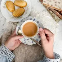 A top-down image of a pair of small, plump, white hands holding a blue and white teacup and saucer. The teacup is full of milky rooibos tea. In the background are some knitting, a couple antique doilies, a plate of madeleines, and some antique books.