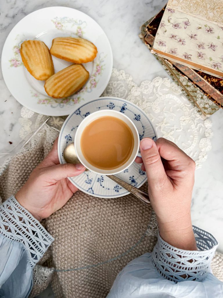 A top-down image of a pair of small, plump, white hands holding a blue and white teacup and saucer. The teacup is full of milky rooibos tea. In the background are some knitting, a couple antique doilies, a plate of madeleines, and some antique books.