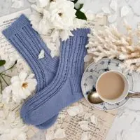 A top down image of a pair of blue handknit socks that take up the left 2/3 of the frame. Their toes are pointing to the left and they're laid slightly cattywumpus on top of each other. They're surrounded by antique paper ephemera, white roses, a hunk of white spindly coral, and a blue and white teacup full of milky tea.