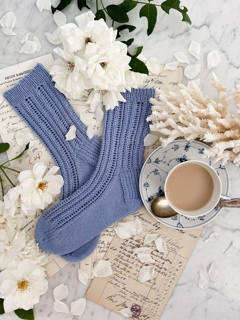 A top down image of a pair of blue handknit socks that take up the left 2/3 of the frame. Their toes are pointing to the left and they're laid slightly cattywumpus on top of each other. They're surrounded by antique paper ephemera, white roses, a hunk of white spindly coral, and a blue and white teacup full of milky tea.
