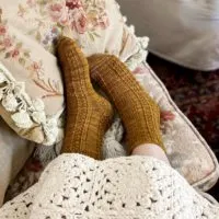 A pair of caramel-colored socks with seed stitch and eyelet panels on two feet, angled slightly to the left. The feet are resting on an embroidered pillow and are peeking out from under a white crocheted blanket.