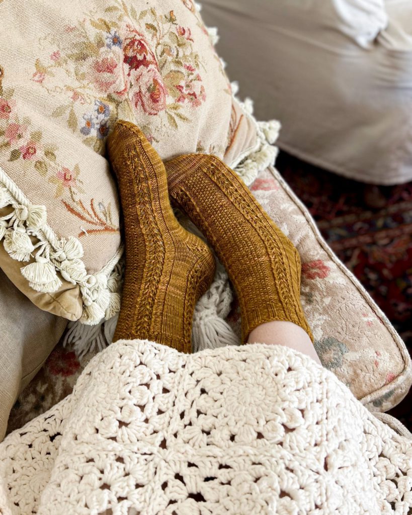 A pair of caramel-colored socks with seed stitch and eyelet panels on two feet, angled slightly to the left. The feet are resting on an embroidered pillow and are peeking out from under a white crocheted blanket.