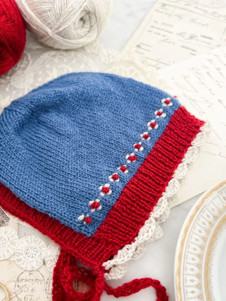 A close-up image of a blue baby bonnet with red ribbing, white scalloped trim, and blue and white roosimine and French knot details. The image is shot from the bottom right corner of the bonnet up toward the crown.