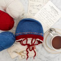 A top-down image of a flatlay on a white marble countertop. In the center is a blue knit baby bonnet. The bonnet has red ribbing around the brim and white scalloped crochet trim. There is white and red roosimine and French knot detailing just above the ribbed brim. The bonnet is surrounded by leftover skeins of yarn, antique paper ephemera, and a gold and white teacup full of tea.