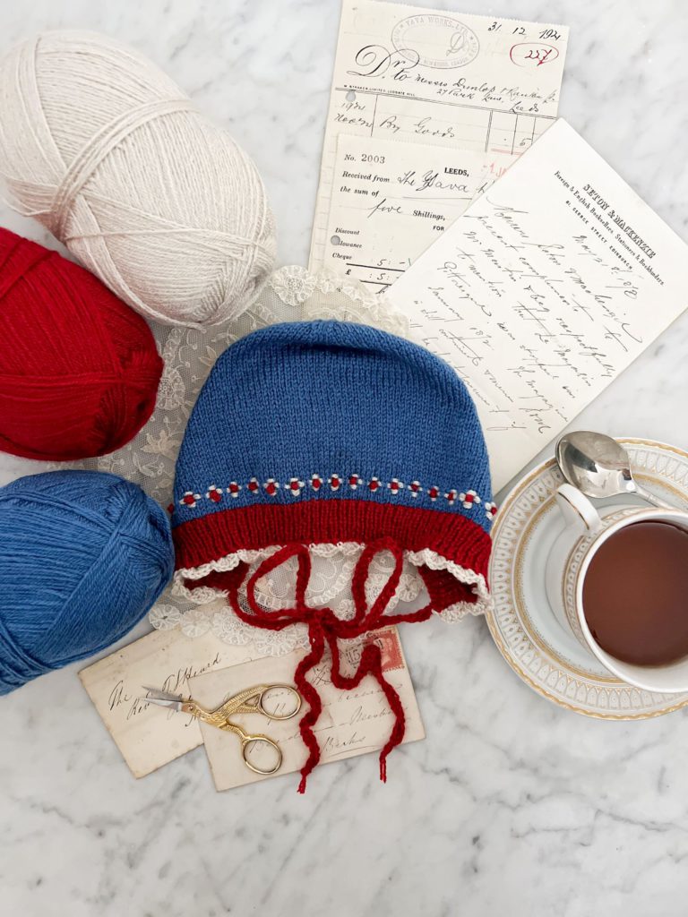 A top-down image of a flatlay on a white marble countertop. In the center is a blue knit baby bonnet. The bonnet has red ribbing around the brim and white scalloped crochet trim. There is white and red roosimine and French knot detailing just above the ribbed brim. The bonnet is surrounded by leftover skeins of yarn, antique paper ephemera, and a gold and white teacup full of tea.