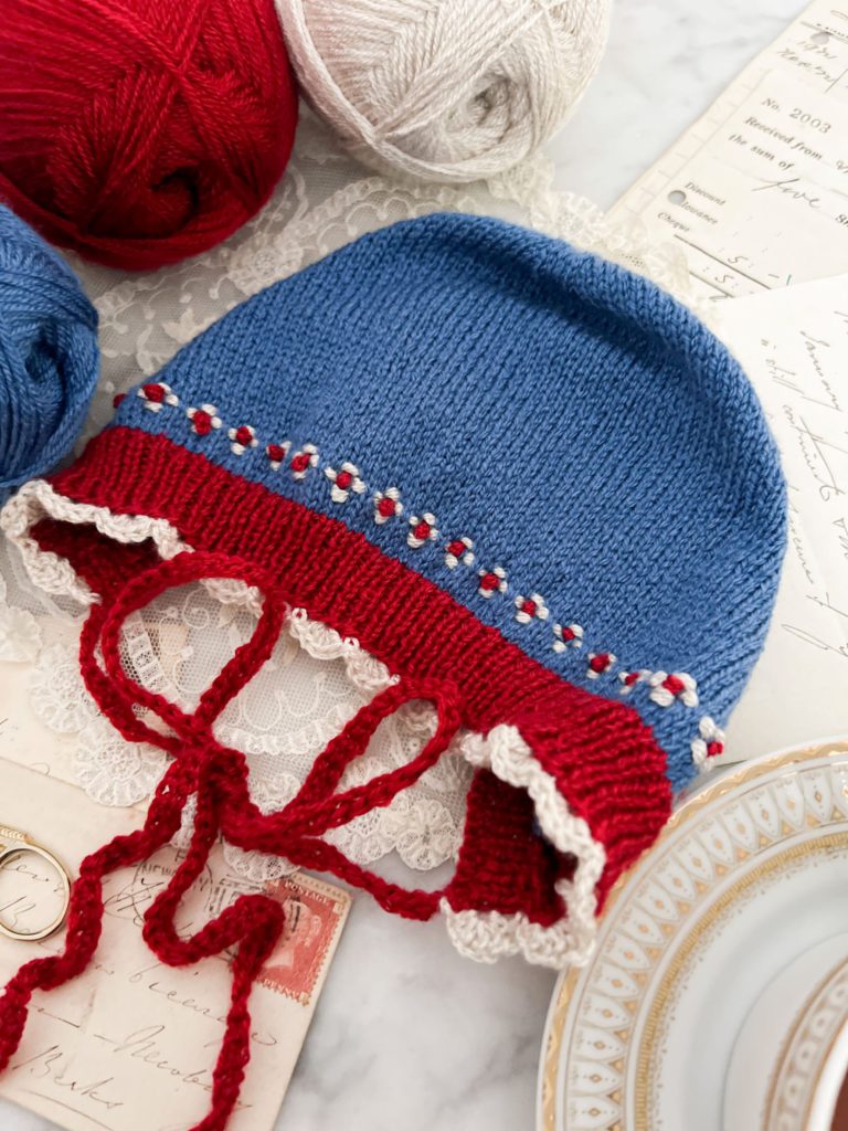 A top-down image of a blue bonnet with red ribbing, white scalloped trim, and red and white roosimine and French knot details. The bonnet is surrounded by balls of yarn and antique paper ephemera.