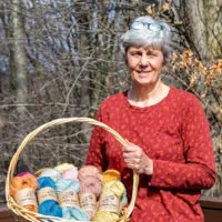 A white woman with short gray hair smiles in the sunshine. She is wearing a red, long-sleeved shirt and has reading glasses perched on top of her head. In her hands is a large basket full of colorful yarn dyed using natural materials.