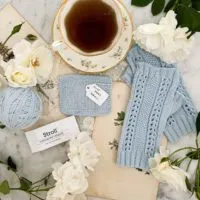 A top-down image of a light blue swatch knit in Knit Picks Stroll, along with a pair of fingerless mitts knit in the same yarn and a ball of leftover yarn. They are surrounded by white roses, antique paper ephemera, and a teacup full of tea.