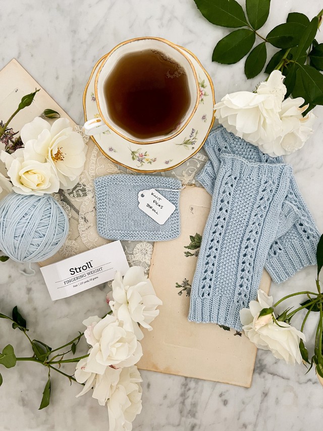 A top-down image of a light blue swatch knit in Knit Picks Stroll, along with a pair of fingerless mitts knit in the same yarn and a ball of leftover yarn. They are surrounded by white roses, antique paper ephemera, and a teacup full of tea.