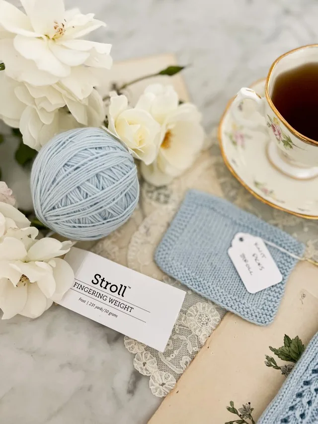 In the foreground is a ball of light blue Knit Picks Stroll, a ball band from that yarn, and some white roses. Blurred slightly in the background are a square swatch of that yarn and a teacup full of tea.