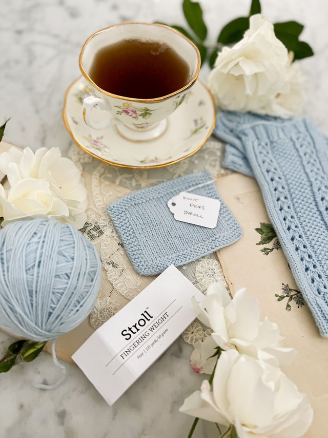 In the foreground is a ball of light blue Knit Picks Stroll and a ball band from the yarn. Slightly blurred behind them are a cup full of tea, a swatch, and a pair of mitts also knit with light blue Knit Picks Stroll.