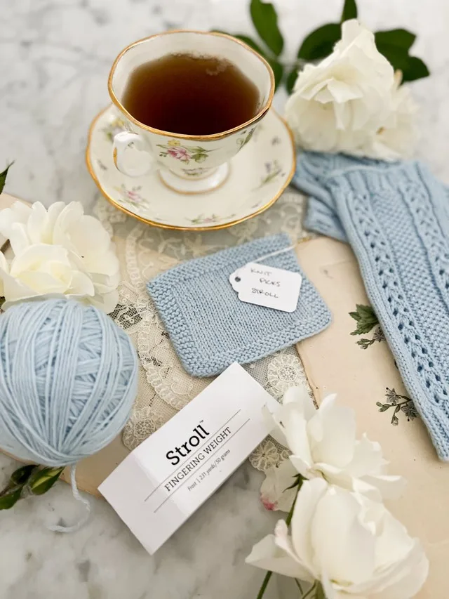 In the foreground is a ball of light blue Knit Picks Stroll and a ball band from the yarn. Slightly blurred behind them are a cup full of tea, a swatch, and a pair of mitts also knit with light blue Knit Picks Stroll.