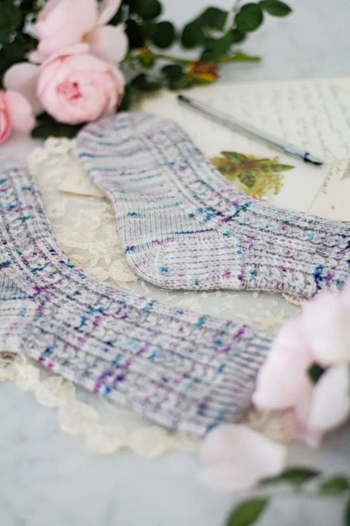 A photo tightly focused on the heel of a gray sock with teal and purple speckles. In the foreground is the matching sock. Blurred in the background is a cluster of pink roses, a silver pen, and some antique paper ephemera.