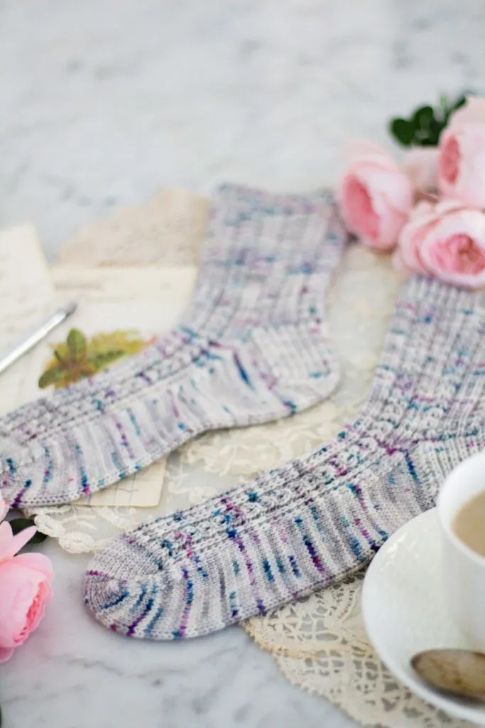 A photo tightly focused on the toe of a gray sock with purple and teal speckles. Blurred in the background are the matching sock and some pink roses.