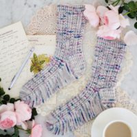 A top-down photo of two handknit socks in gray with teal and purple speckles. They feature columns of coin lace and slipped stitches for lots of texture. The toes are pointing to the left. They're surrounded by pink roses, antique paper ephemera, and a white teacup and saucer full of milky tea.