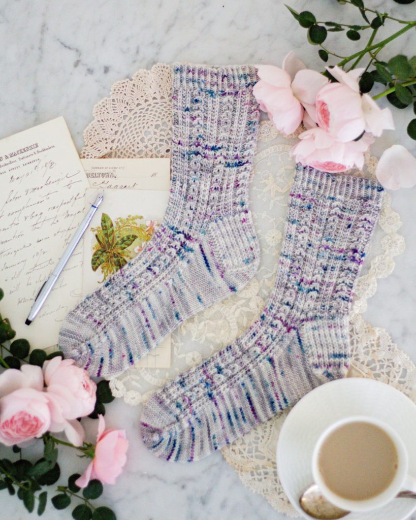 A top-down photo of two handknit socks in gray with teal and purple speckles. They feature columns of coin lace and slipped stitches for lots of texture. The toes are pointing to the left. They're surrounded by pink roses, antique paper ephemera, and a white teacup and saucer full of milky tea.