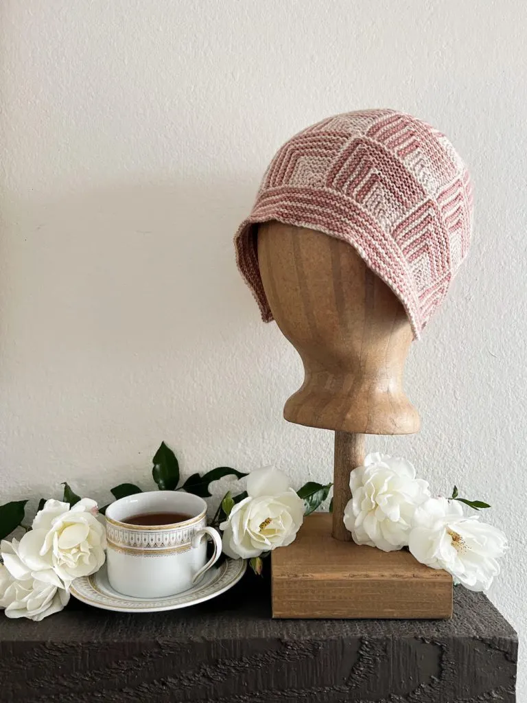 A cloche-shaped knit hat made of pink and cream mitered squares sits on top of a brown head form. The head form is on a dark brown shelf and surrounded by white roses. A white and gold teacup sits on the shelf next to it.