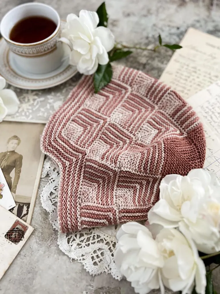 A close-up image of a hat knit from pink and cream mitered squares. It's surrounded by white roses and a teacup full of tea.