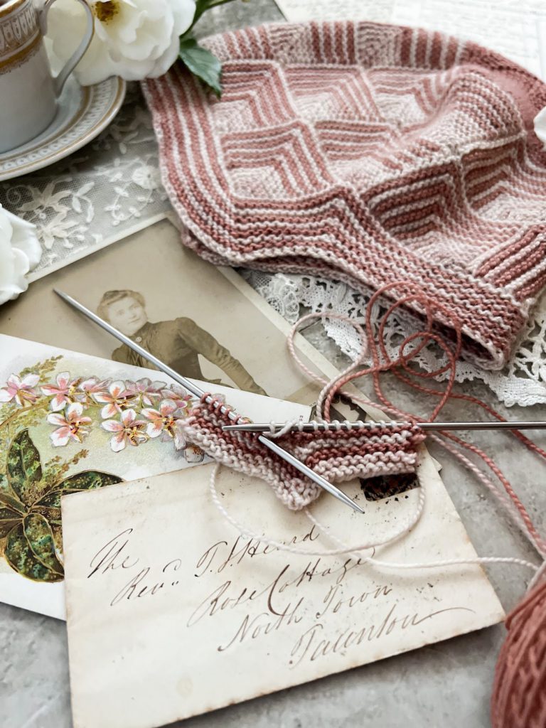 A close-up image focusing on a pink and cream mitered square in progress on a pair of tiny steel needles. It sits on top of several pieces of antique paper ephemera. In the background is a hat knit from mitered squares in the same colors.