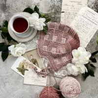 A pink and cream hat composed entirely of mitered squares is laid flat on a textured gray surface. It's surrounded by a gold and white teacup full of tea, white roses, antique paper ephemera, two cakes of leftover yarn in the same colors as the hat, and a mitered square in progress on two tiny steel needles.