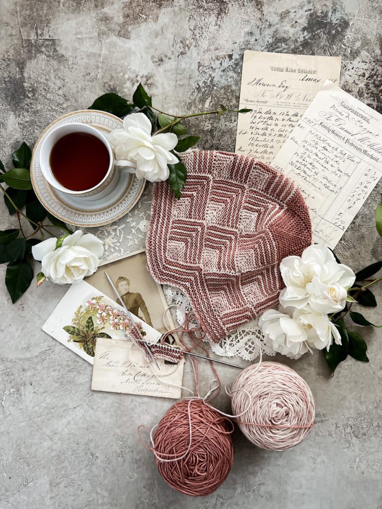 A pink and cream hat composed entirely of mitered squares is laid flat on a textured gray surface. It's surrounded by a gold and white teacup full of tea, white roses, antique paper ephemera, two cakes of leftover yarn in the same colors as the hat, and a mitered square in progress on two tiny steel needles.