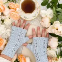 A woman's small, plump hands are splayed slightly on a white countertop. They're wearing a pair of light blue fingerless mitts with panels of lace and seed stitch down the back of the hands. Around them are lots of peach and cream roses. A teacup full of tea sits at the top of the photo.
