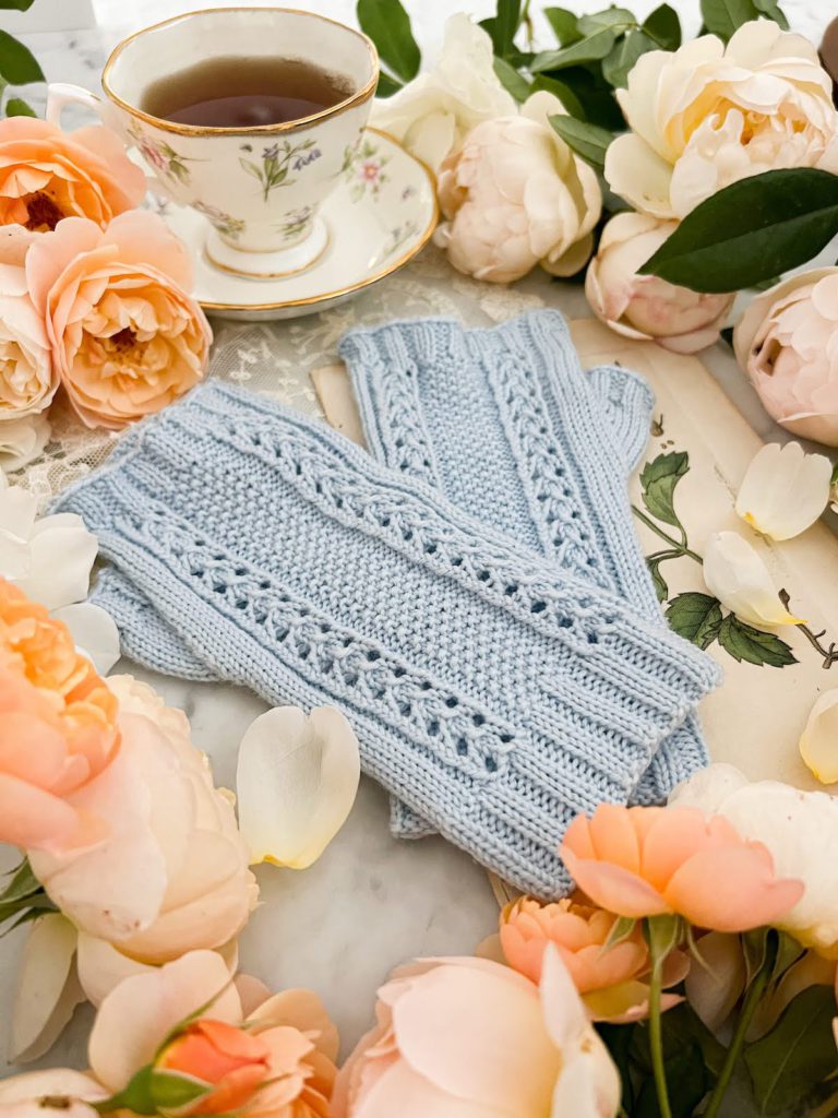 A pair of light blue fingerless mitts is laid flat on a countertop surrounded by peach and cream roses. The mitts are photographed from the wrist cuff angled upward toward the fingertips.