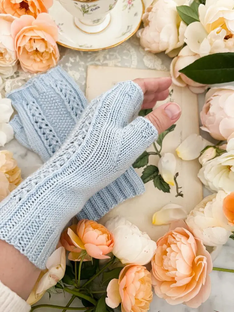 A close up on the left thumb gusset of the Arroyo Verde Mitts, a pair of light blue fingerless mitts. In the background are lots of peach and cream roses and an antique botanical print.
