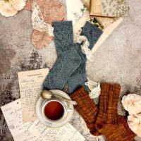 Three pairs of knit socks (pink, blue, and caramel) are laid out on a flat surface in a diagonal from top left to bottom right. They're surrounded by white roses, antique books, a teacup full of chamomile, and antique paper ephemera.