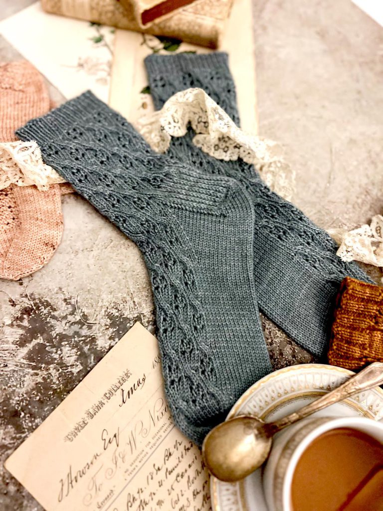 A pair of blue hand-knit socks is laid flat on a textured surface. They're surrounded by other pairs of socks (almost entirely cropped out), a white and gold teacup, some antique lace, and some antique paper ephemera.