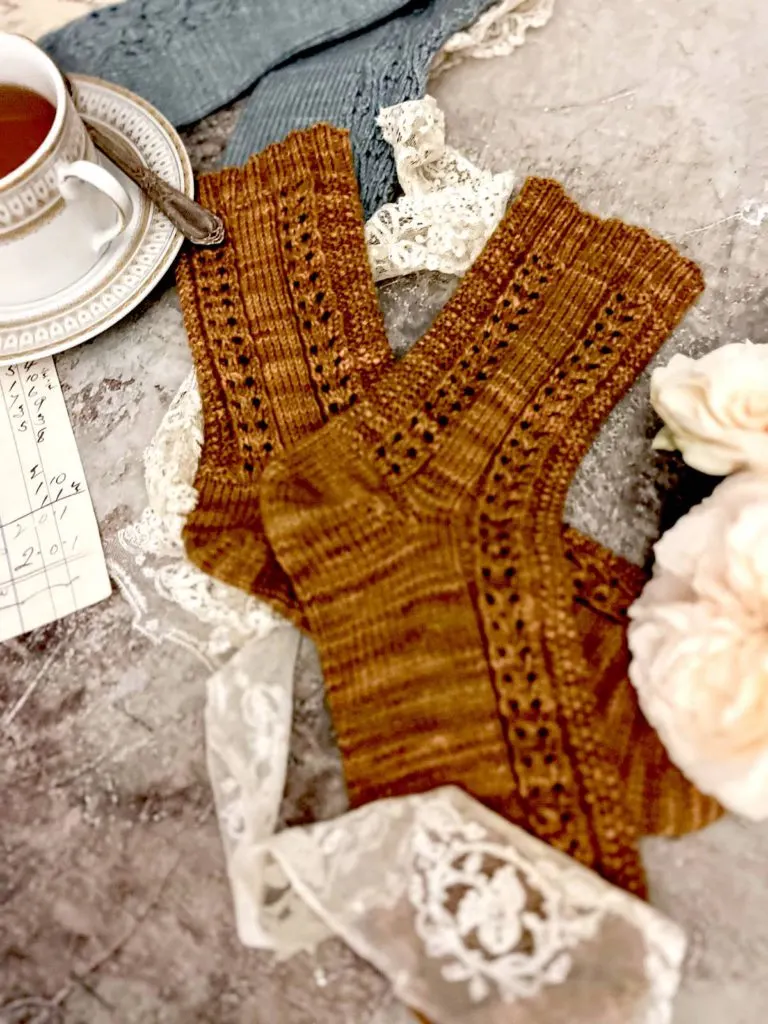 A pair of caramel-colored knit socks is laid flat on a textured surface. The foreground is slightly blurred, with the focus on the highly textured leg-portion in the middle of the photo. In the background is a pair of blue socks and a white and gold teacup.
