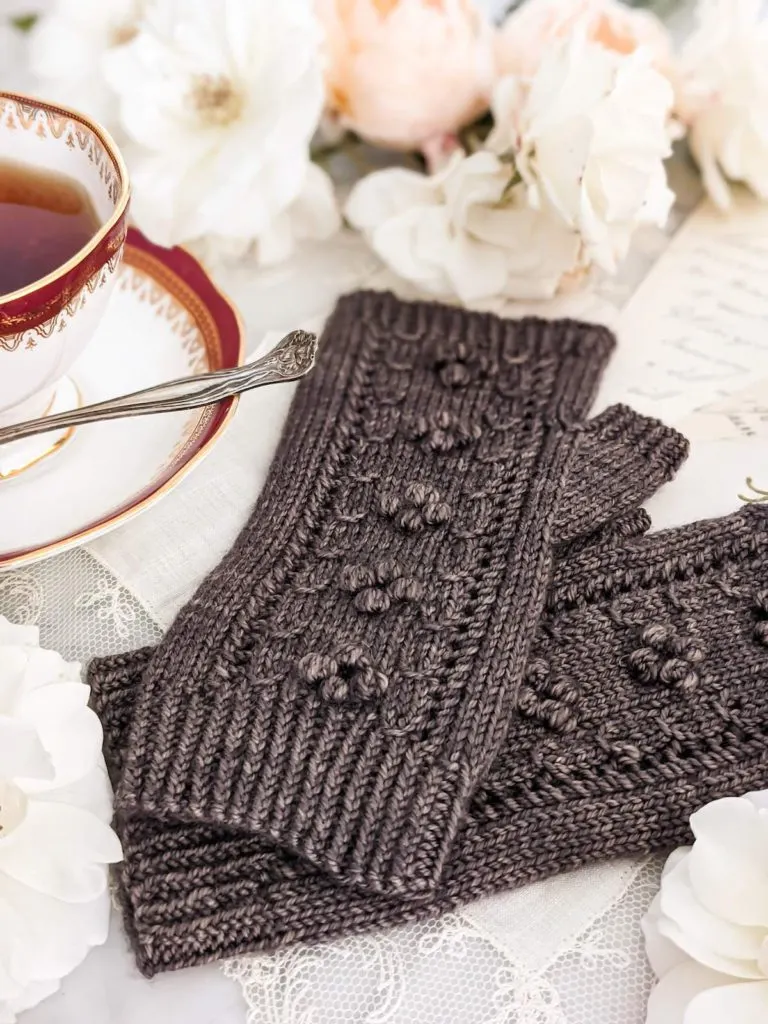 A pair of brown fingerless mitts with little bobbles on the back is laid out on an antique handkerchief trimmed with lace. In the background are some peach and cream roses.