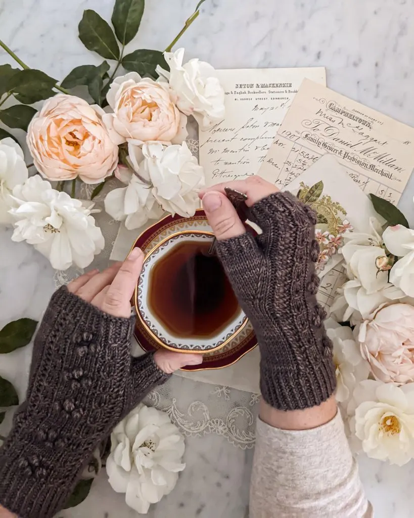 A woman's small, plump hands stir a cup of tea while wearing brown fingerless mitts. In the background are peach and cream roses and some antique paper ephemera.