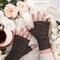 A woman's small, plump hands are splayed slightly on a white countertop. They're wearing a pair of brown fingerless mitts with panels of coin lace and little bobbles down the back of the hands. Around them are some peach and cream roses and antique paper ephemera. A teacup full of tea sits to the left, slightly cropped out.