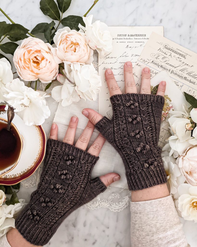 A woman's small, plump hands are splayed slightly on a white countertop. They're wearing a pair of brown fingerless mitts with panels of coin lace and little bobbles down the back of the hands. Around them are some peach and cream roses and antique paper ephemera. A teacup full of tea sits to the left, slightly cropped out.