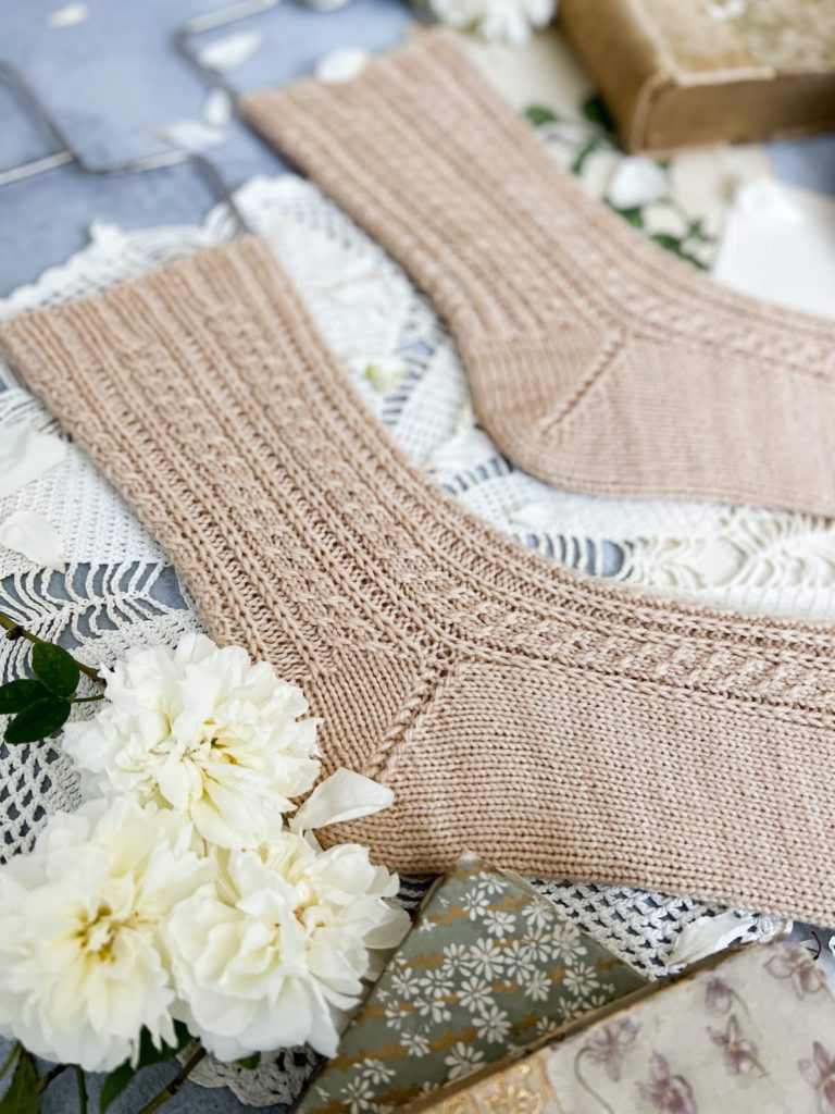 A close-up on the heel of a pale pink sock with tiny cables. The heel is a boomerang-style heel made with German short rows. In the background is the sock's mate. In the foreground are some tiny white roses.