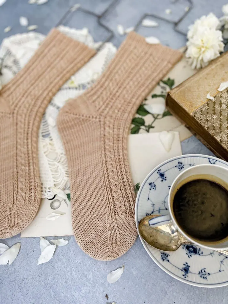 In focus in the foreground is the toe of a light pink sock with tiny cables. Next to it is a blue and white teacup full of espresso. The legs of the socks extend into the background and blur as they move farther way from the foreground.