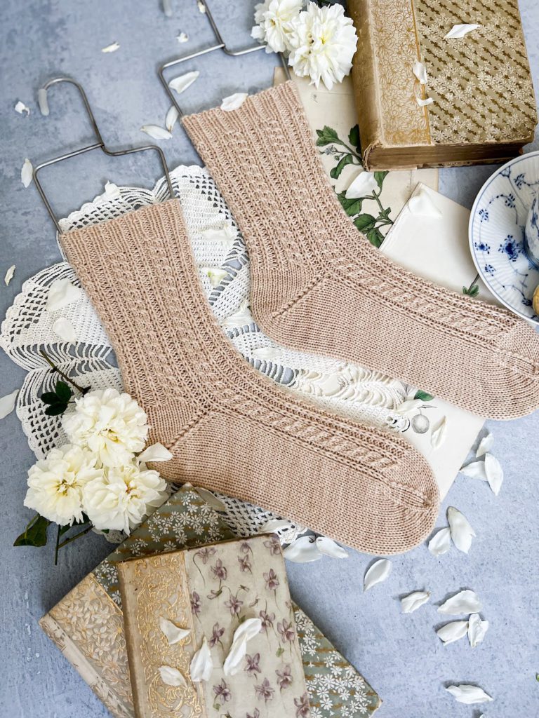 A pair of pale pink socks with tiny cables and a short-row heel are laid out flat on a gray surface. The toes are pointing to the right. The socks are surrounded by antique paper ephemera, old books, and white roses.