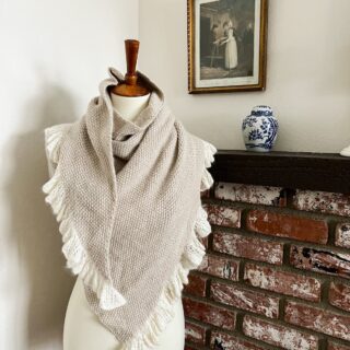 The Grandmillennial Shawl is draped around a white dressmaker's form. It's a seed stitch shawl in an asymmetrical triangle shape that has a ruffled edge. The ruffles are made from a creamy silk-mohair blend, while the body of the yarn is knit in a luxurious DK-weight cahmere-merino yarn from Purl Soho.