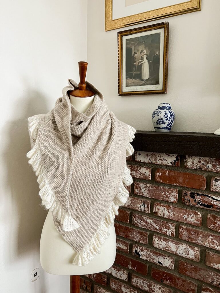 The Grandmillennial Shawl is draped around a white dressmaker's form. It's a seed stitch shawl in an asymmetrical triangle shape that has a ruffled edge. The ruffles are made from a creamy silk-mohair blend, while the body of the yarn is knit in a luxurious DK-weight cahmere-merino yarn from Purl Soho.