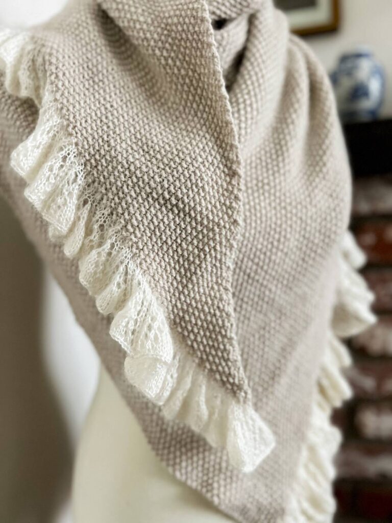 A close-up on the narrowest point of the Grandmillennial Shawl, a gray, seed stitch shawl that's draped around a dressmaker's form. This photo shows how the fabric drapes and how dramatically the ruffle expands from the edge.