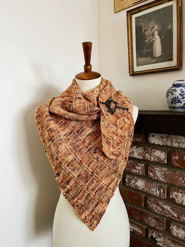 A speckled orange shawl is draped around a white dressmaker's form and secured with a shawl pin.