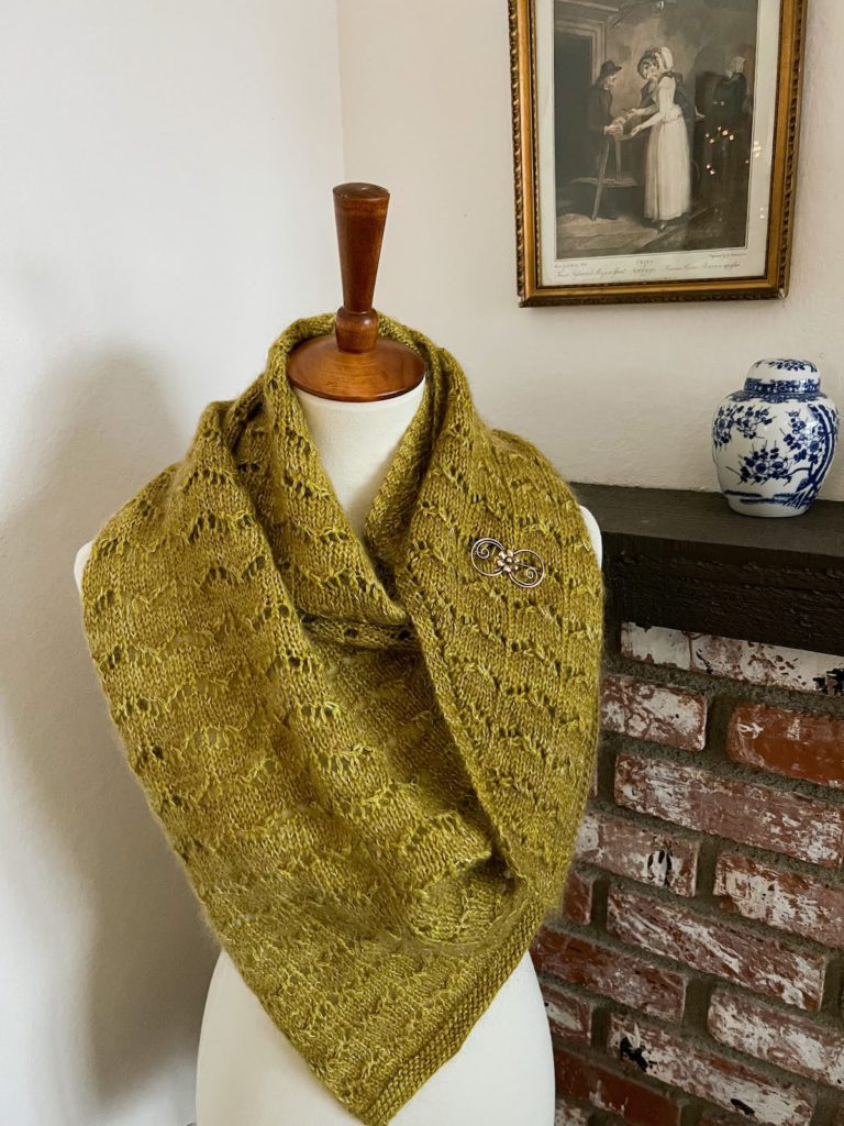 A green, lacy shawl is draped around a white dressmaker's form and secured with a scrolling brass brooch.