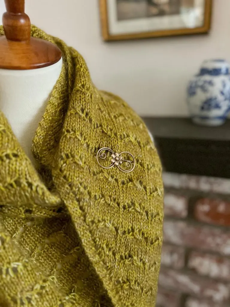A close-up of a swirling brass brooch securing a green lacy shawl.