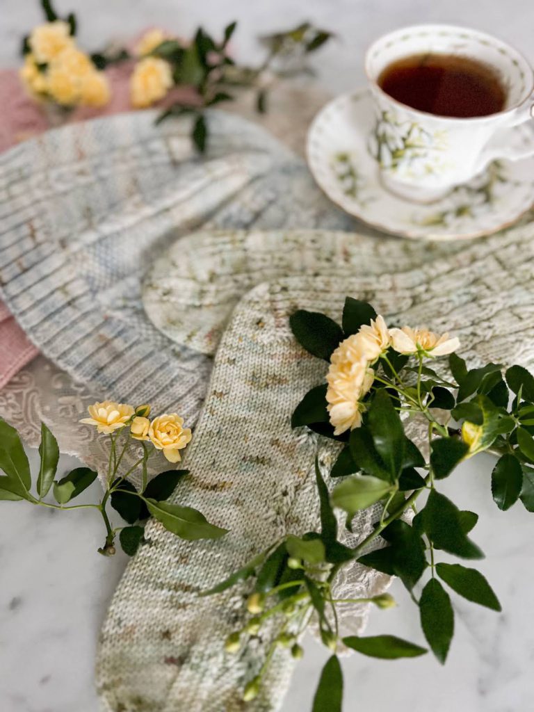 A pair of green socks is in the foreground of the image. Several tiny yellow roses sit on top of the socks. Blurred in the background are a blue hat, pink socks, more roses, and a white teacup full of tea.