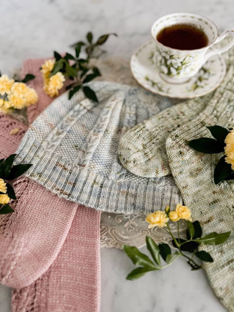 The middle of the image focuses on a pale blue hat and the toes of a pair of green socks. Slightly blurred in the foreground are a pair of pink sock toes. Slightly blurred in the background is a white teacup full of tea. There are tiny yellow roses scattered throughout the image.