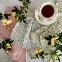 A top-down image of a pair of pink socks, a light blue hat, and a pair of green socks all laid flat on a white marble countertop. They're surrounded by tiny yellow roses and a white teacup full of tea.